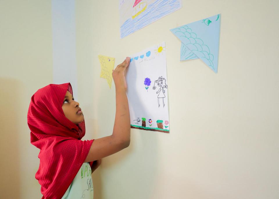 A young girl tapes up a drawing inside a UNICEF-supported Child-Friendly Space at the Karkar bus station in Aswan Egypt, established to provide psychosocial support displaced children and families fleeing the conflict in Sudan.