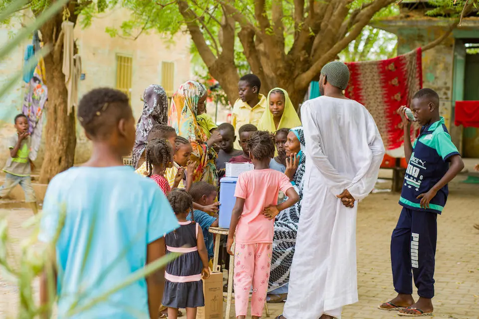 A UNICEF-supported vaccinator at Gezirat Al-Feel Health Center in Sudan speaks to mothers and caregivers who have brought their children for vaccination at a shelter for displaced people.