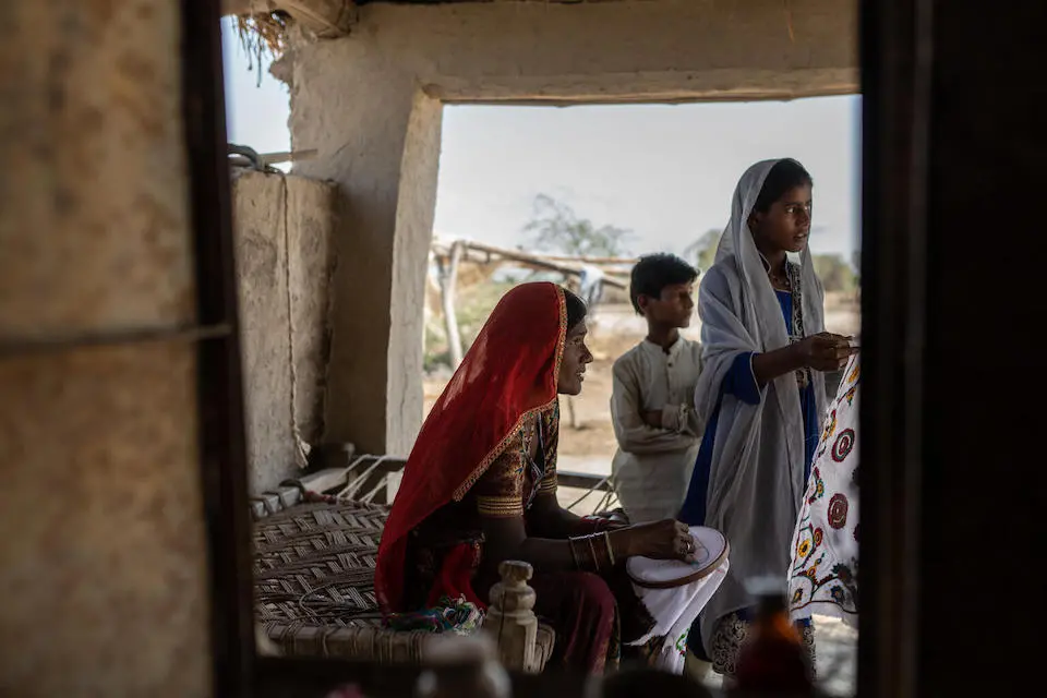 A mother in flood-devastated Pakistan sews textiles in the shade of her porch as her children stand nearby.