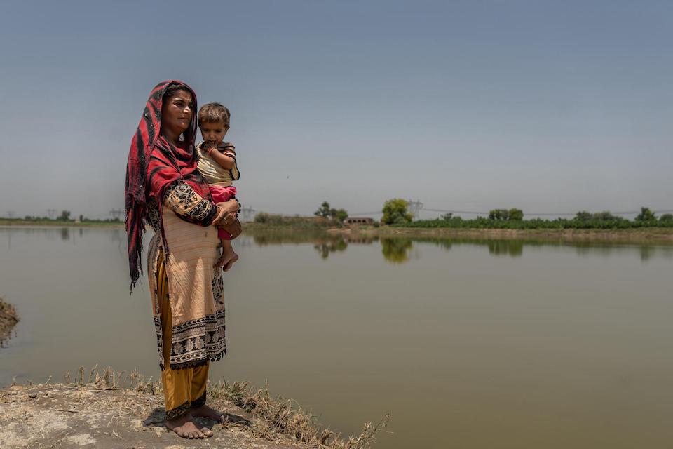 In May 2023, a mother stands holding her 3-year-old daughter near stagnant water in the village of Balocho Zardari, Shaheed Benazirabad, an area of Sindh, Pakistan where mothers and children are still suffering the effects of the catastrophic flooding that hit the country during the 2022 monsoon season.