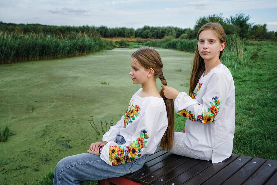 On Sept. 9, 2023, on the banks of the river in Horenka village near Kyiv, Ukraine, 14-year-old Nelya gazes into the distance while her 13-year-old sister Lilya braids her hair. 