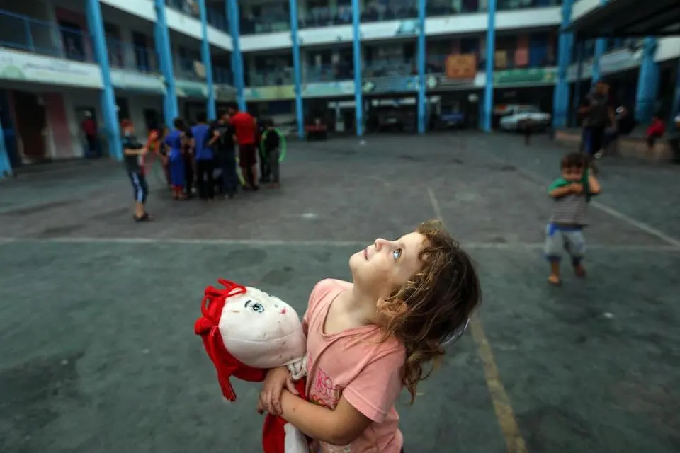 Aya, a 5-year-old girl clutching her doll to ease her fear, gazing at Gaza's sky filled with warplanes from inside an UNRWA school in the Gaza Strip.