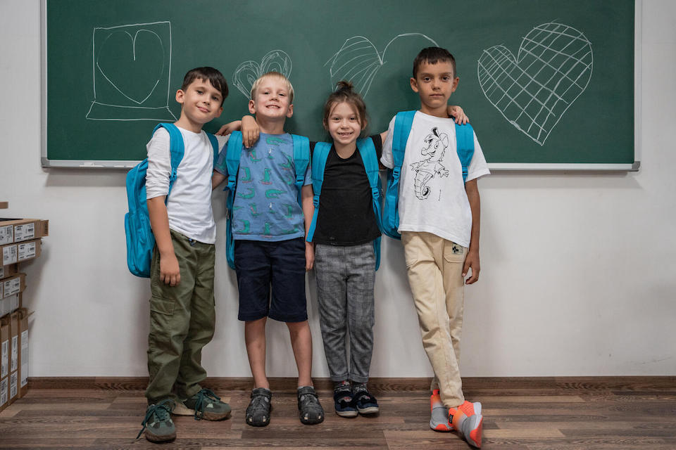 Four young children from Ukraine who are refugees from the war stand in front of a chalkboard in a Romanian classroom with their UNICEF backpacks.