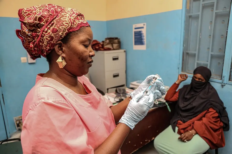 A midwife at a health center in Mali prepares a tetanus vaccine as a seated woman looks on.