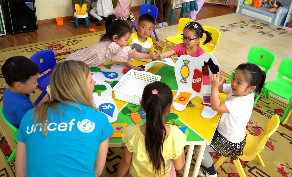 A UNICEF USA staff member sits at a table in a kindergarten classroom with children during a classroom activity about nutrition and dental health during a program visit to Ulaanbaatar, Mongolia, May 2024.