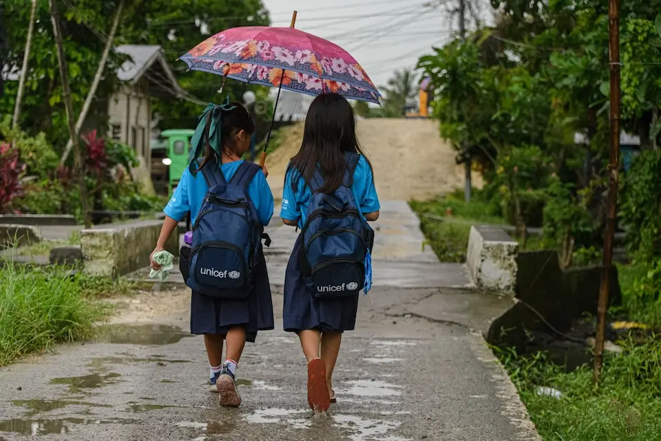 In Barangay Loob, Gainza, Camarines Sur, Philippines, two girls share with blue UNICEF backpacks walk to school sharing an umbrella.