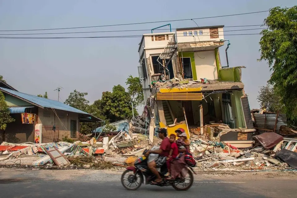 On March 31, 2025, a man rides a motorcycle with children and a woman on board, passing by a collapsed building in Sagaing, Myanmar following a 7.7 magnitude earthquake on March 28.