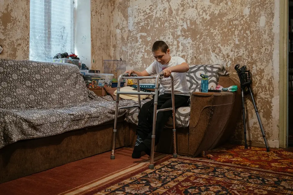 In his living room in eastern Ukraine, a boy badly injured by an explosive remnant of war tries to stand using a walker. 