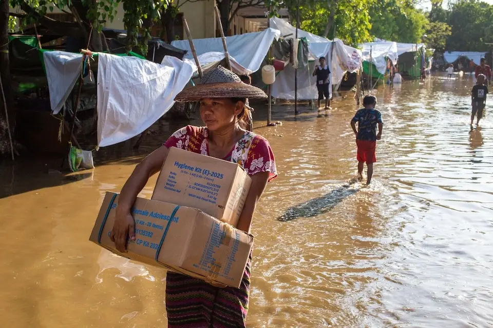 On April 16, 2025, a woman carries humanitarian relief supplies inside the flooded camp compound at BEHS-33 Temporary Camp during a distribution by UNICEF Myanmar.