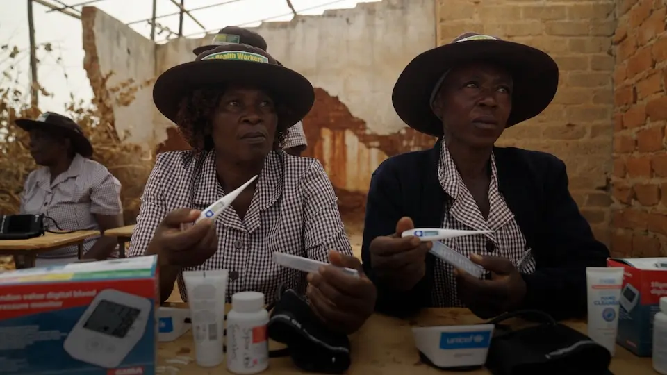 Two women wearing hats and matching dresses sit side by side at a table holding up digital thermometers