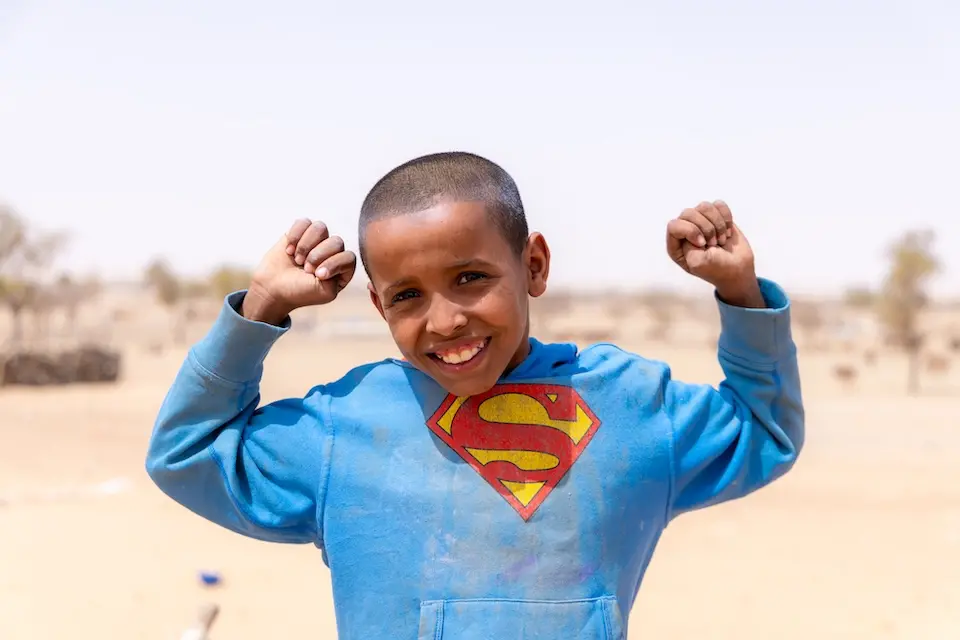 A boy in Mauritania wearing a Superman T-shirt smiles with arms raised.