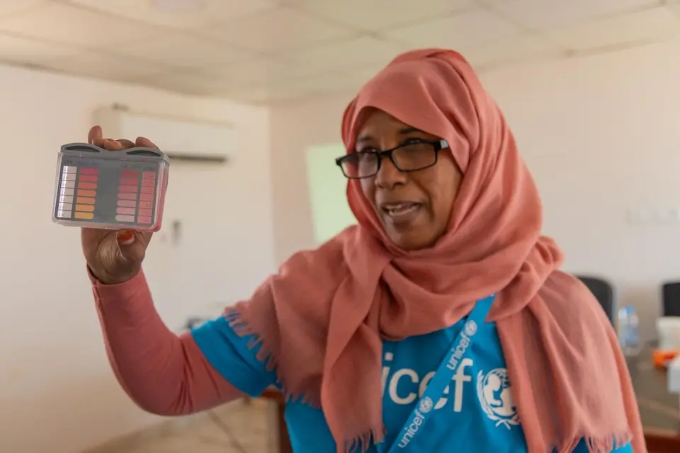 During a cholera outbreak in Sudan, a woman wearing a UNICEF shirt and headscarf holds a device used to check water quality. 
