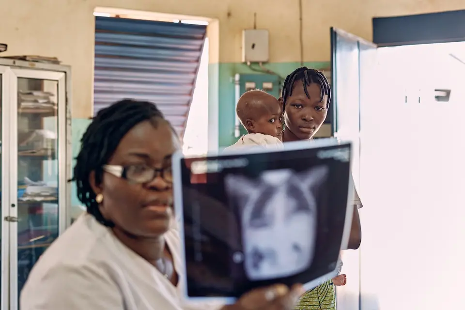 Woman wearing lab coat seated in foreground examines a chest x-ray, standing behind her, a young woman holds an infant girl