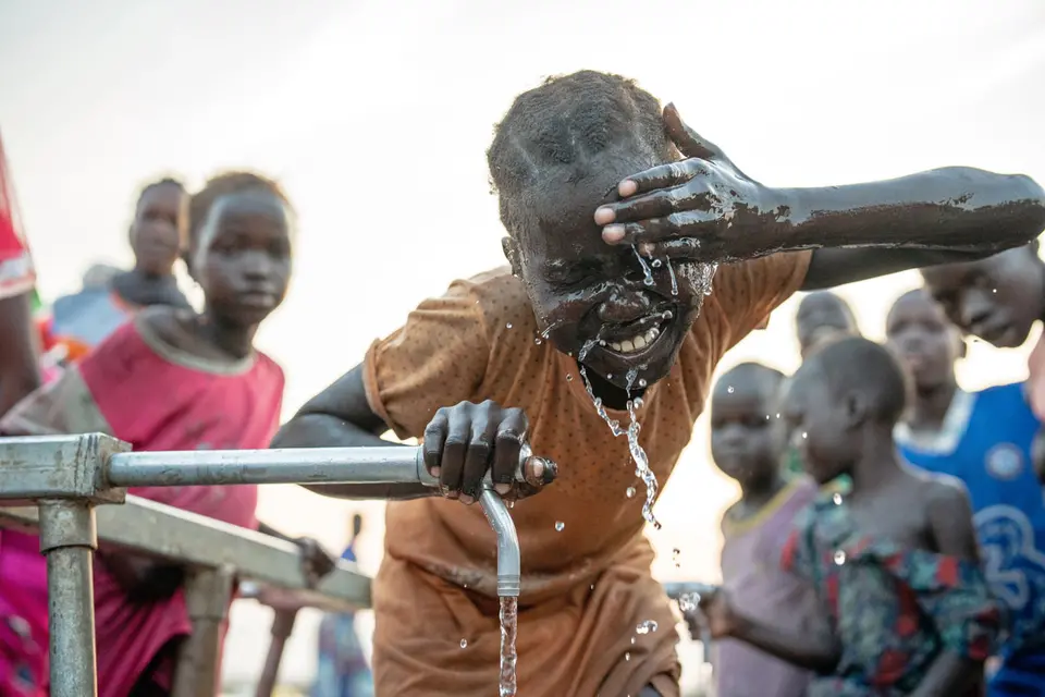A child joyfully splashes water on their face from a hand pump, surrounded by other children. The scene appears to be in a rural or developing area, highlighting access to clean water.