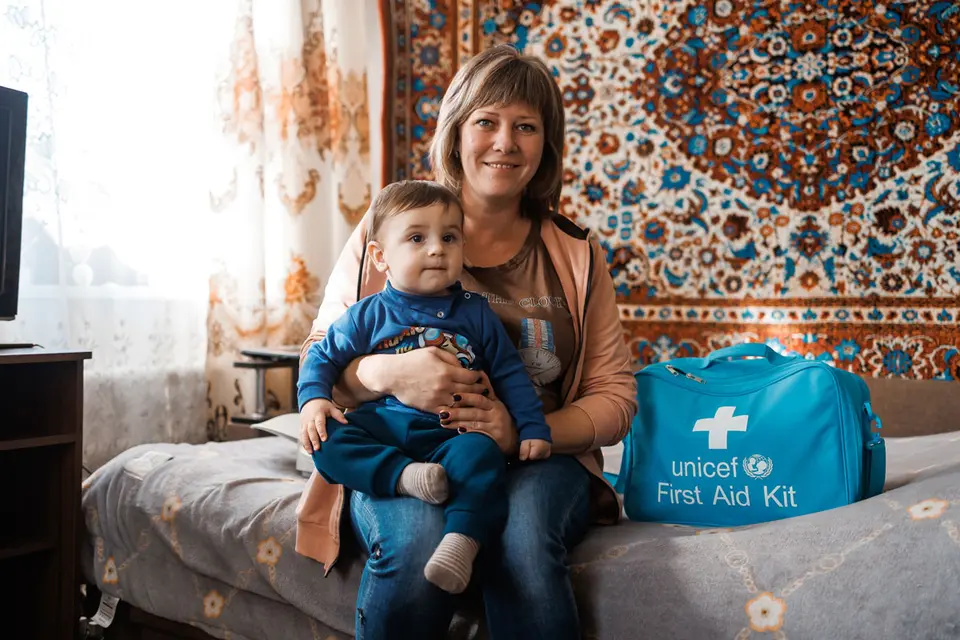 A smiling woman sits on a bed holding a toddler in her lap. Beside them is a bright blue UNICEF First Aid Kit bag. The room is warmly lit with patterned curtains and a decorative carpet hanging on the wall.