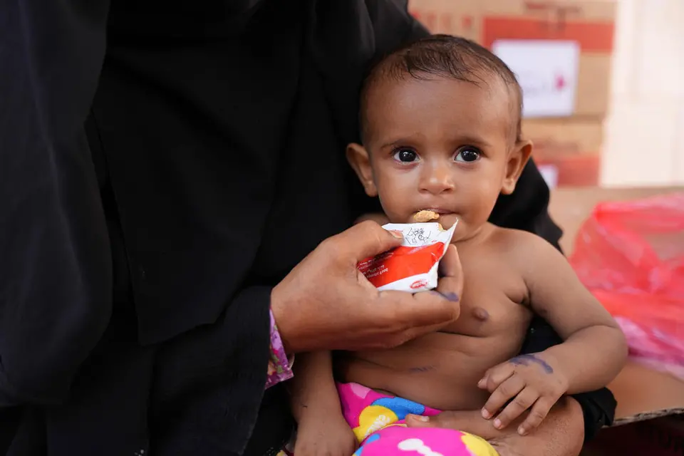 A baby sits on an adult's lap while being fed a therapeutic food paste from a packet. The baby looks directly at the camera, with cardboard boxes in the background suggesting an aid distribution setting.