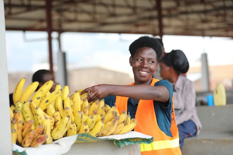 Smiling woman wearing a safety vest, standing behind a box of bananas and reaching for a banana.