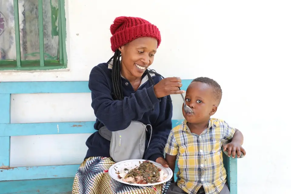 Woman sitting with plate on lap feeding small child standing next to her from plate