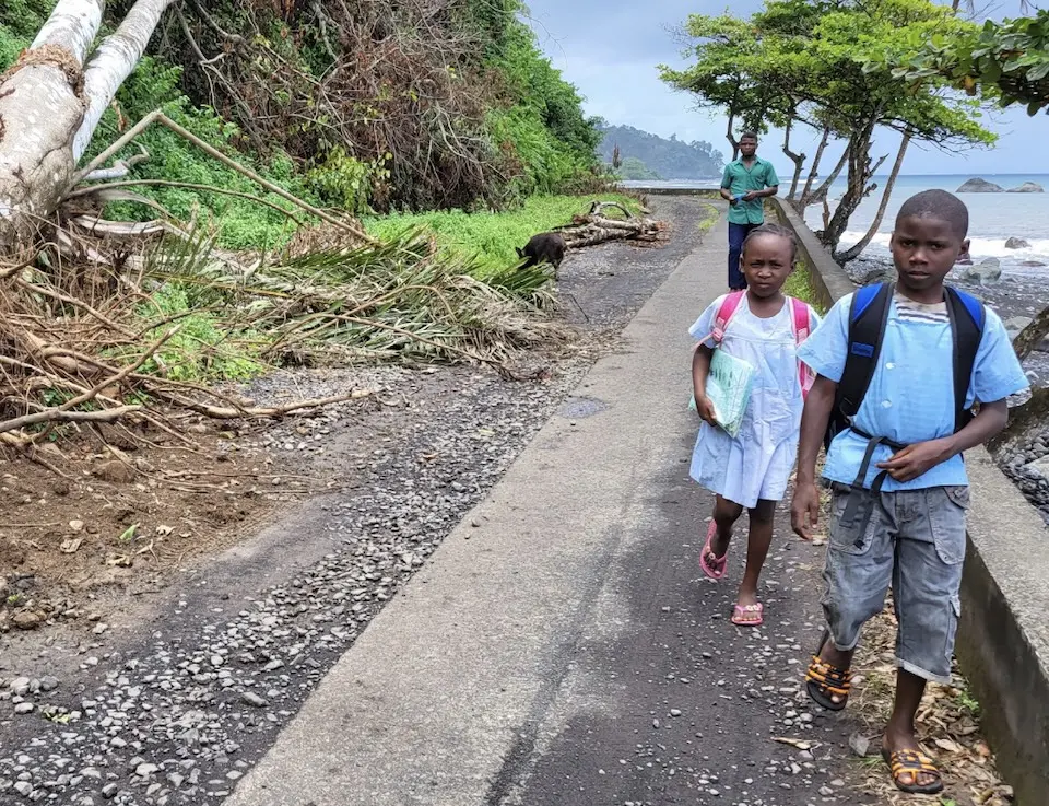 A girl and a boy carrying backpacks walk back from school along a path showing environmental damage in a northern coastal part of São Tomé and Príncipe.