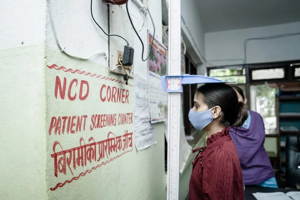 Young girl being measured for height, facing wall with writing "NCD Corner. Patient Screen Center"