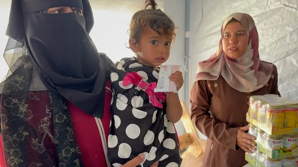On May 7, 2025, at Al Farooq Camp in Khan Younis, Gaza, a woman holds a small child who grips a packet of ready-to-use therapeutic food (RUTF).