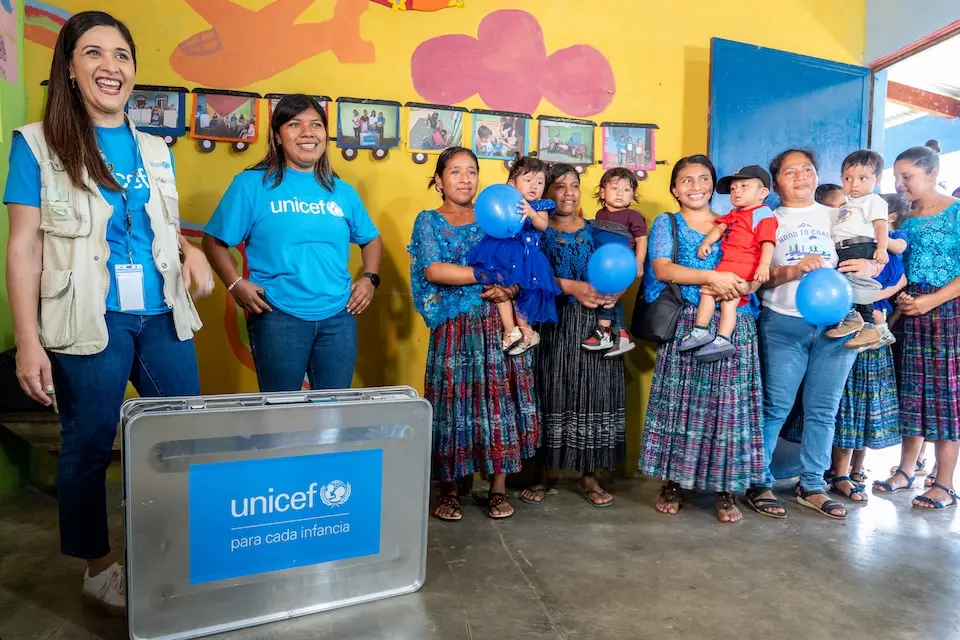 Woman in UNICEF vest and blue t-shirt stands behind a metal case with "UNICEF para cada infancia" printed on the side, to the left other UNICEF workers and child development center parents hold children and balloons and smile