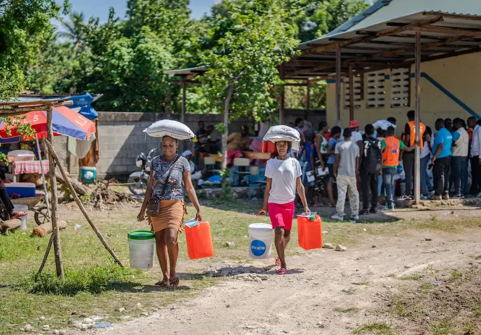 In southern Haiti, two women leave a a UNICEF hurricane aid distribution point carrying water buckets and other items.