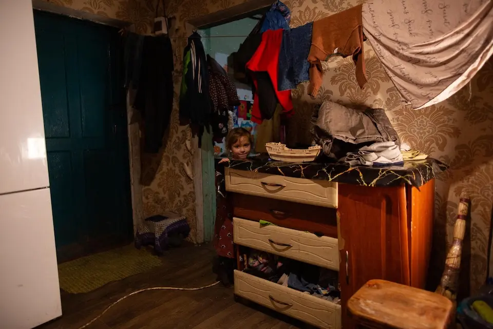 A child peeks out from behind a dresser, with clothes drying overhead, in the family's temporary house in eastern Ukraine.