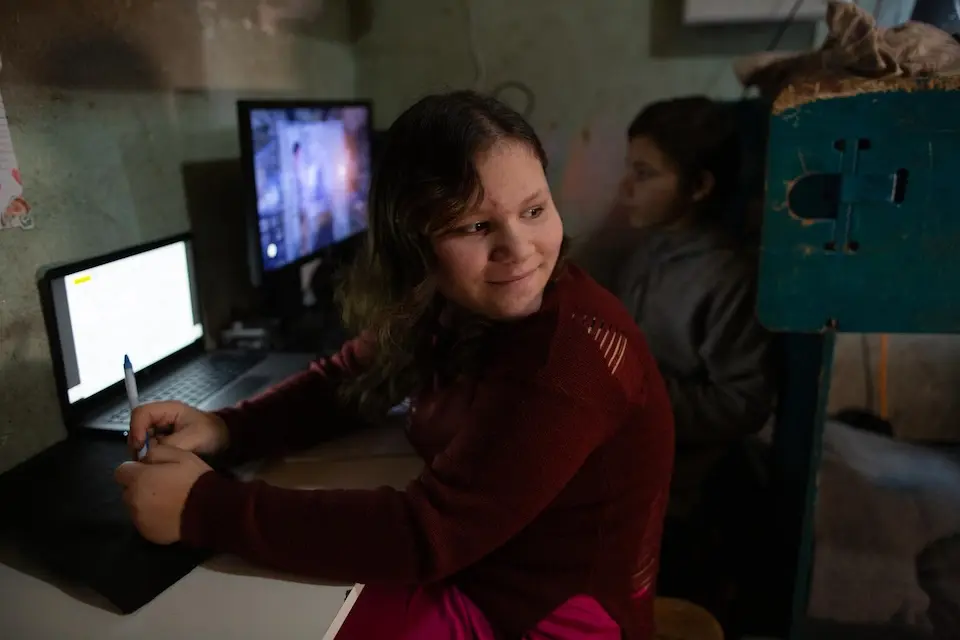 A girl sits at a small table doing her schoolwork on a computer while her brother plays an educational game in their temporary home in Staryi Saltiv, Kharkiv region, Ukraine.