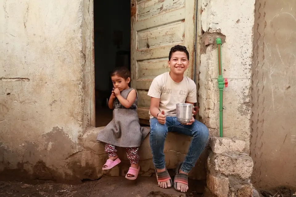 A young boy and girl sitting in a doorway. Boy smiles at camera holding a jug of water in his left hand and his right thumb up.