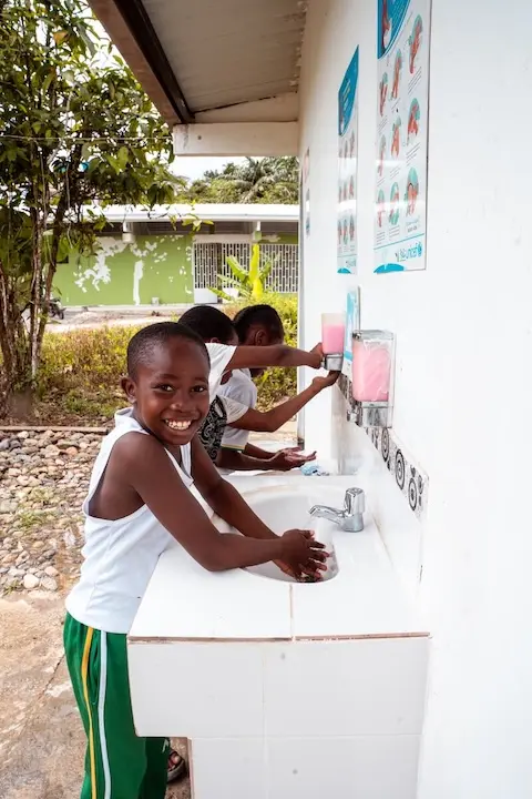 Young boy facing camera while washing his hands at a row of outdoor sinks. Behind him are several other children washing their hands.