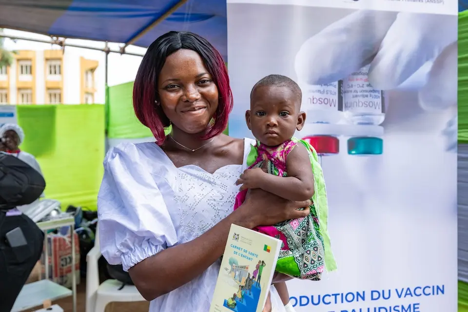 A mother in Benin holding her infant child in her arms while learning about malaria vaccination.