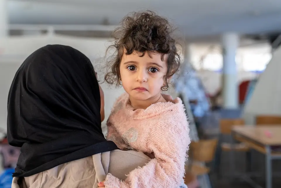 On March 17, 2026, a 2-year-old girl is held by an older girl at a school in Beirut being used as a shelter for families seeking safety.