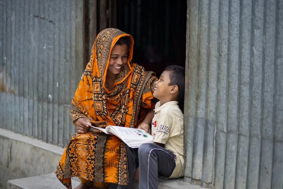 Woman in orange head covering smiles at young boy sitting next to her, holding a book