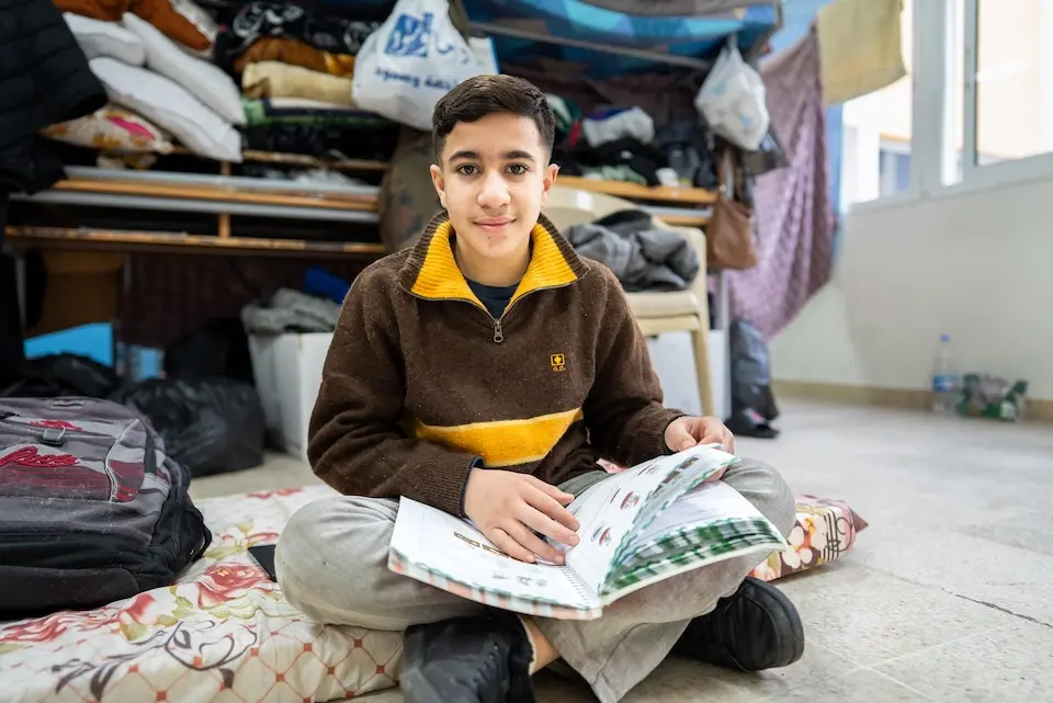 A boy sits on the floor with a workbook open on his lap in Lebanon.