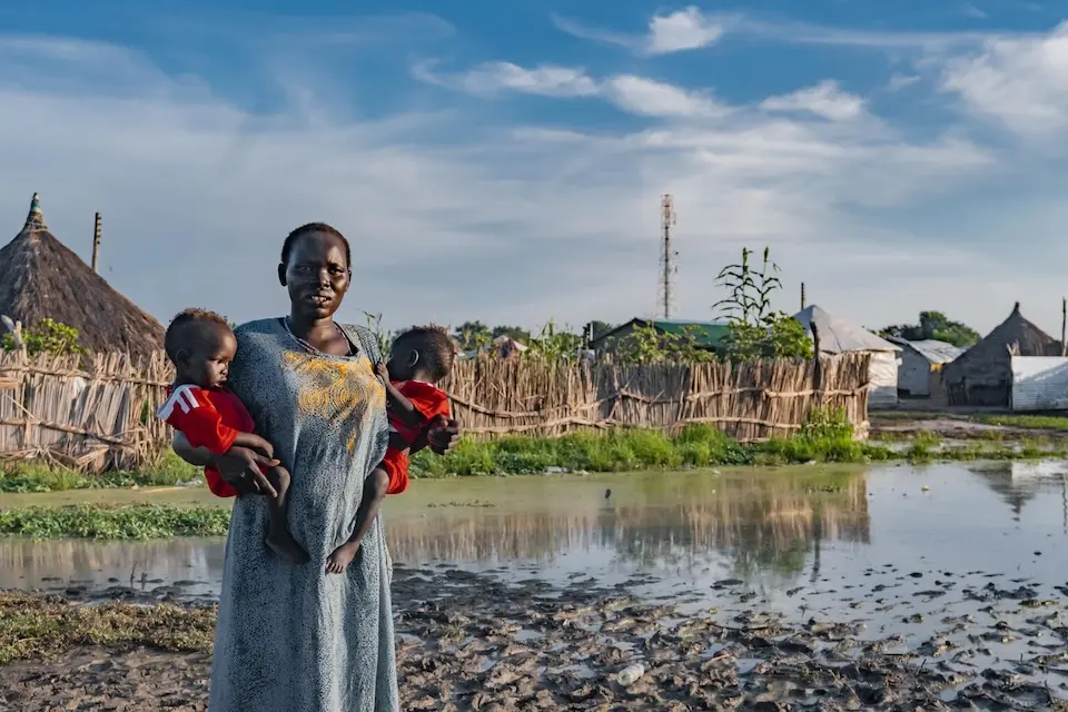 A mother holds two small children on both hips by a flooded riverband in South Sudan.