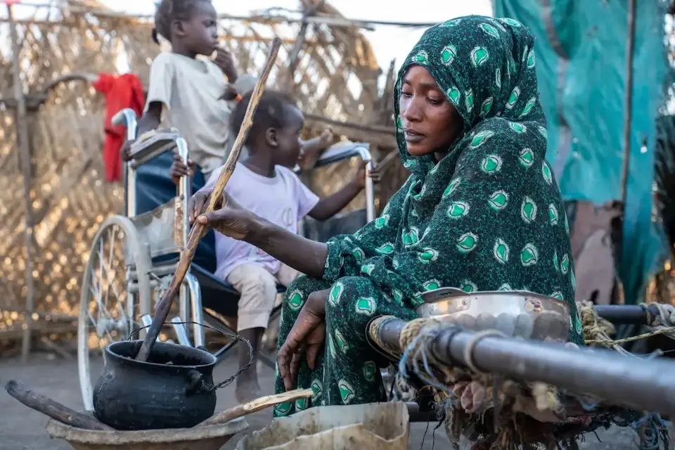 Seated on the ground, a mother stirs a pot of food while one of her children sits in her wheelchair in rural Gedaref, Sudan. 