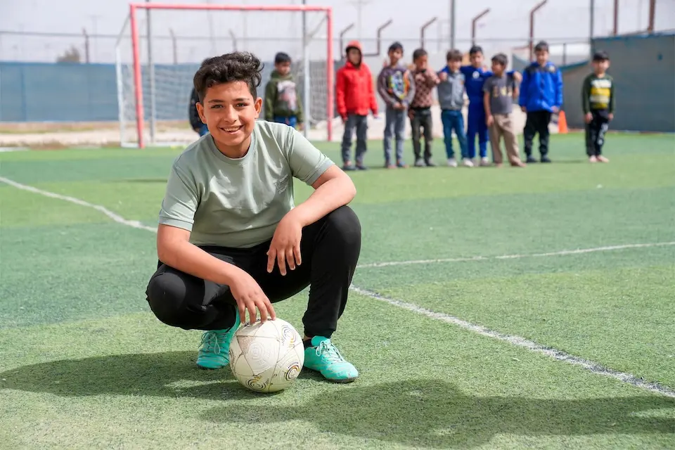 A Syrian refugee boy with a soccer ball kneels on a soccer field inside a UNICEF-supported camp in Jordan.