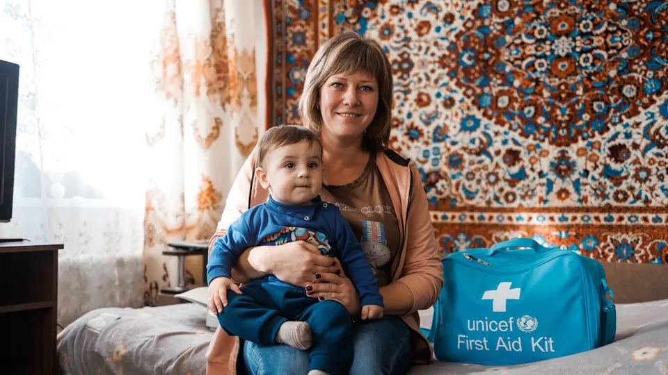 A smiling woman sits on a bed holding a toddler in her lap. Beside them is a bright blue UNICEF First Aid Kit bag. The room is warmly lit with patterned curtains and a decorative carpet hanging on the wall.