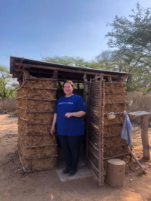 Women in blue tee shirt reading "Baxter Gives Back" poses beside latrine built of branches and stone.