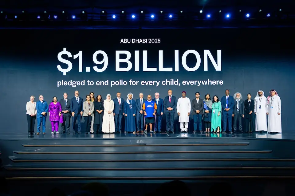 Large group of adults standing on a stage under a banner that states the group has pledged 1.9 billion to end polio