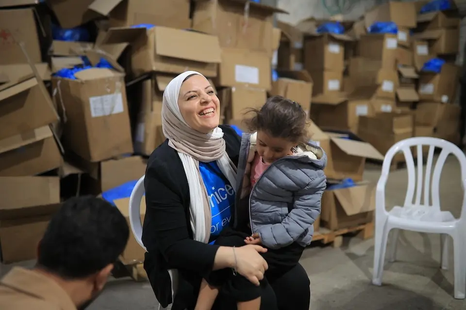 In the Gaza Strip on Oct. 6, 2026, a UNICEF staff member holds a child on her lap in a room full of boxes of winter clothes for children. 