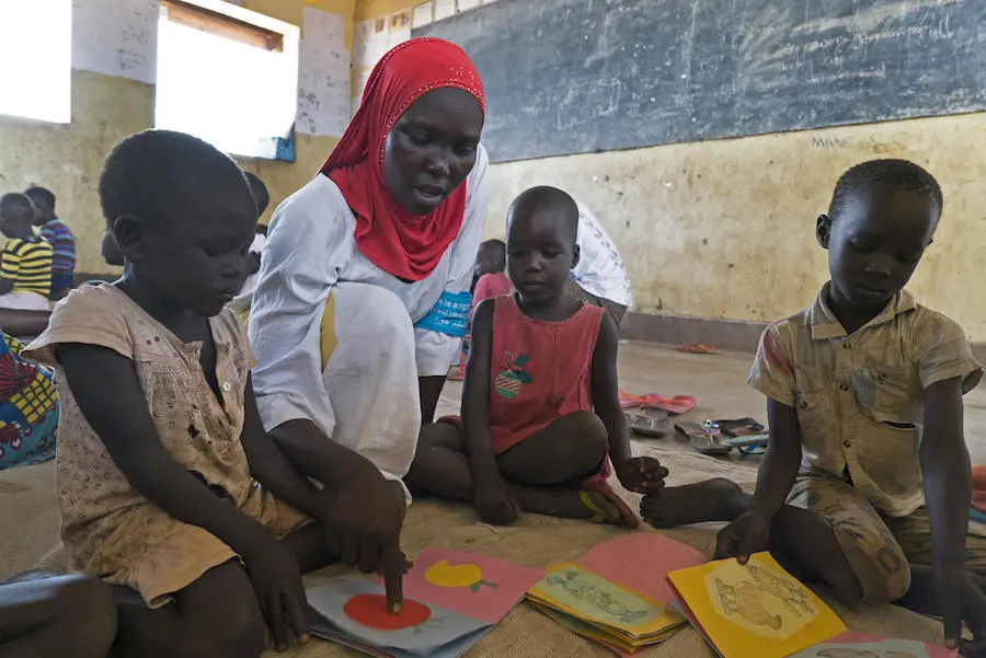 Ugandan caregiver Dalla Alli, 28, shows locally made cardboard books to South Sudanese refugee children in Bidi Bidi refugee settlement. Ugandan caregiver Dalla Alli, 28, shows locally made cardboard books to South Sudanese refugee children in Bidi Bidi refugee settlement.