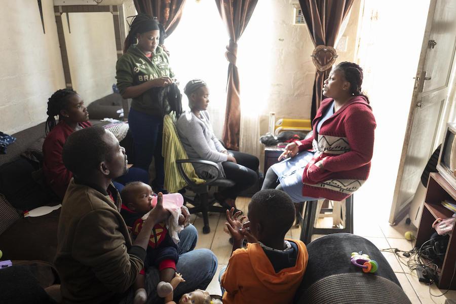Dieu-Merci Matala, 44, bottle-feeds baby Grace while his wife, Bibiche, standing, braids a client's hair in the family's apartment in Maitland, Cape Town, South Africa in May 2019.