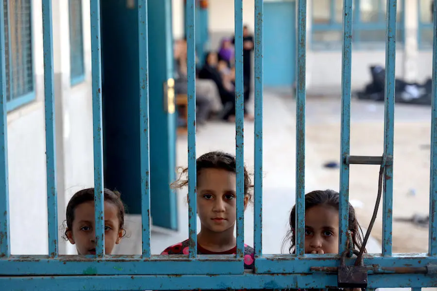 Three girls peeking outside a gate at one of 29 schools used as temporary shelter during in Gaza in May 2021. Three girls peeking outside a gate at one of 29 schools used as temporary shelter in Gaza in May 2021.