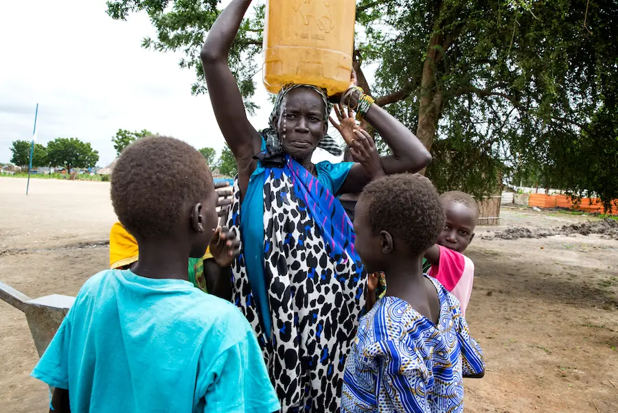 Angelina Nyakuma is surrounded by her children as she carries a jerry can of safe drinking water, drawn from a water pump at the Machakos Primary School, Bentiu, South Sudan, July 2017. Angelina Nyakuma is surrounded by her children as she carries a jerry can of safe drinking water, drawn from a water pump at the Machakos Primary School, Bentiu, South Sudan, July 2017.