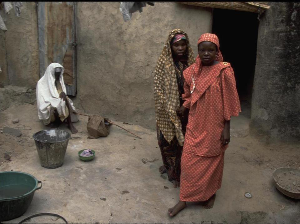 In 1997 a girl, far right, guides her mother, who lost her sight from river blindness, past cooking utensils and debris in an alley where a neighbor sits, outside their home in Kaduna, Nigeria. 