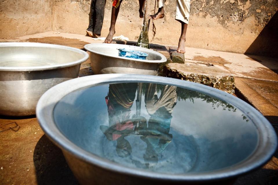 Children fill containers with water from a communal foot-activated pump, in the village of Kiendi-Walogo, Zanzan Region. A reflection of two children is in the closest water bowl.