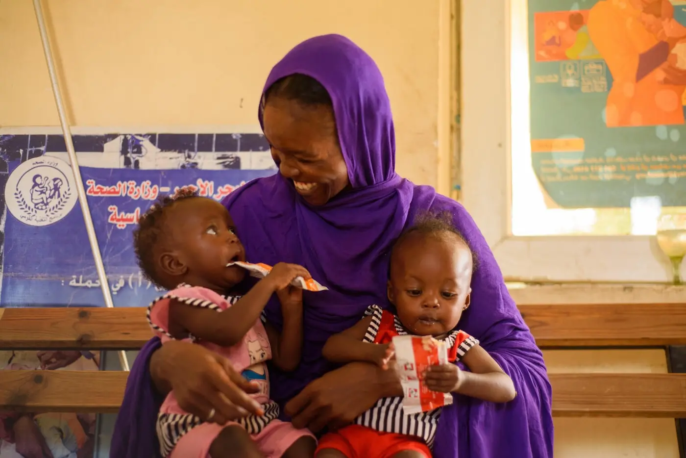 At a UNICEF-supported health facility in Port Sudan, a mother holds her twin daughters in her lap as both consume packets of Ready-to-Use Therapeutic Food, a peanut paste UNICEF supplies to support the recovery of acutely malnourished children.