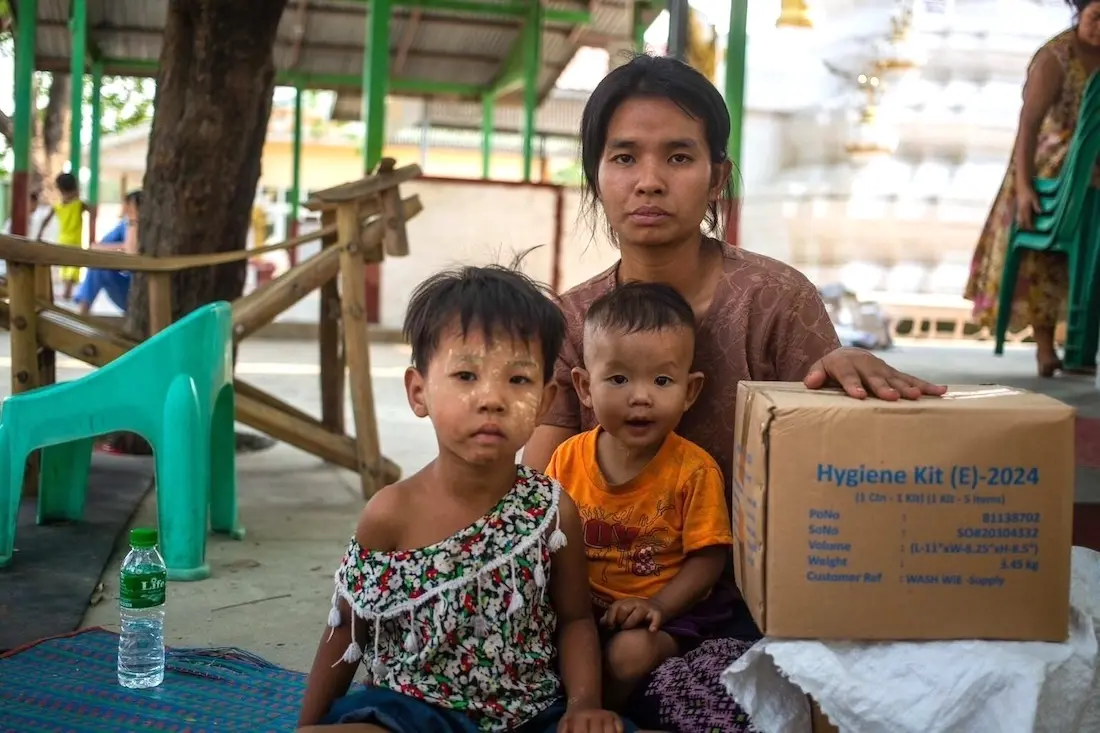 On March 31, 2025, a woman with two small children poses for a photograph with a box of UNICEF hygiene kits distributed to earthquake survivors in the Sagaing region of Myanmar.